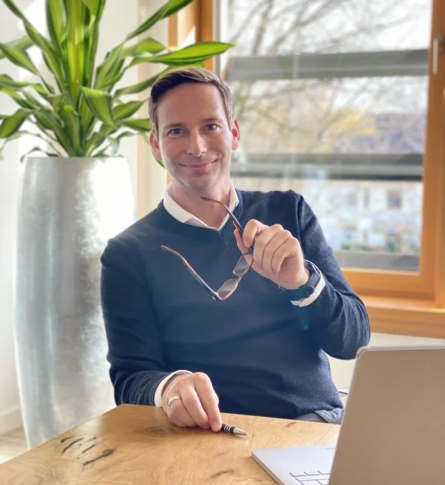 Andre Achtermeier at his desk with a laptop, holding his glasses and smiling at the camera